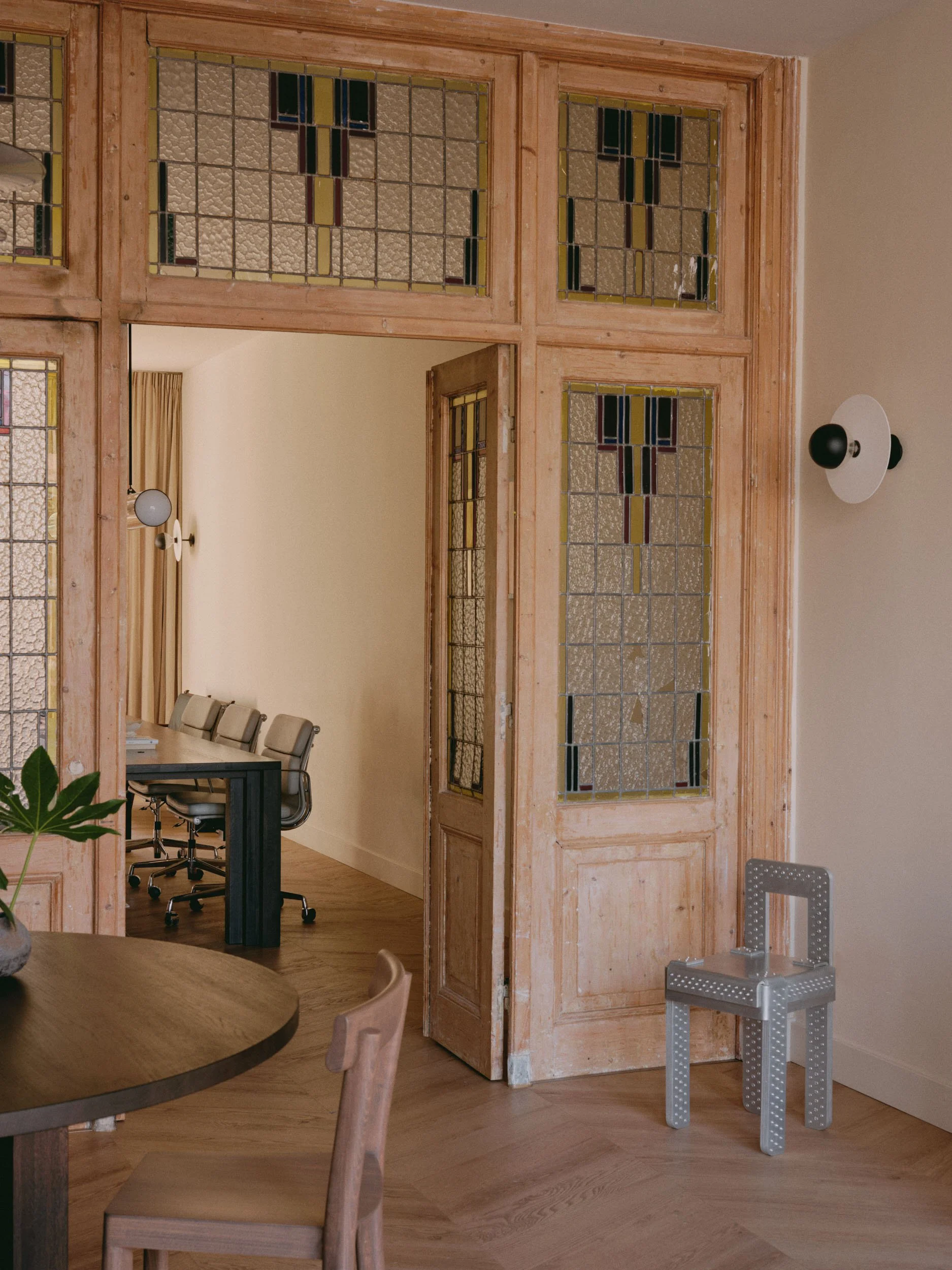 Interior of an apartment or office with a wooden doorframe and stained glass panels, a round wooden table with a chair, a small grey chair with holes, a potted plant, and a wall-mounted light fixture.