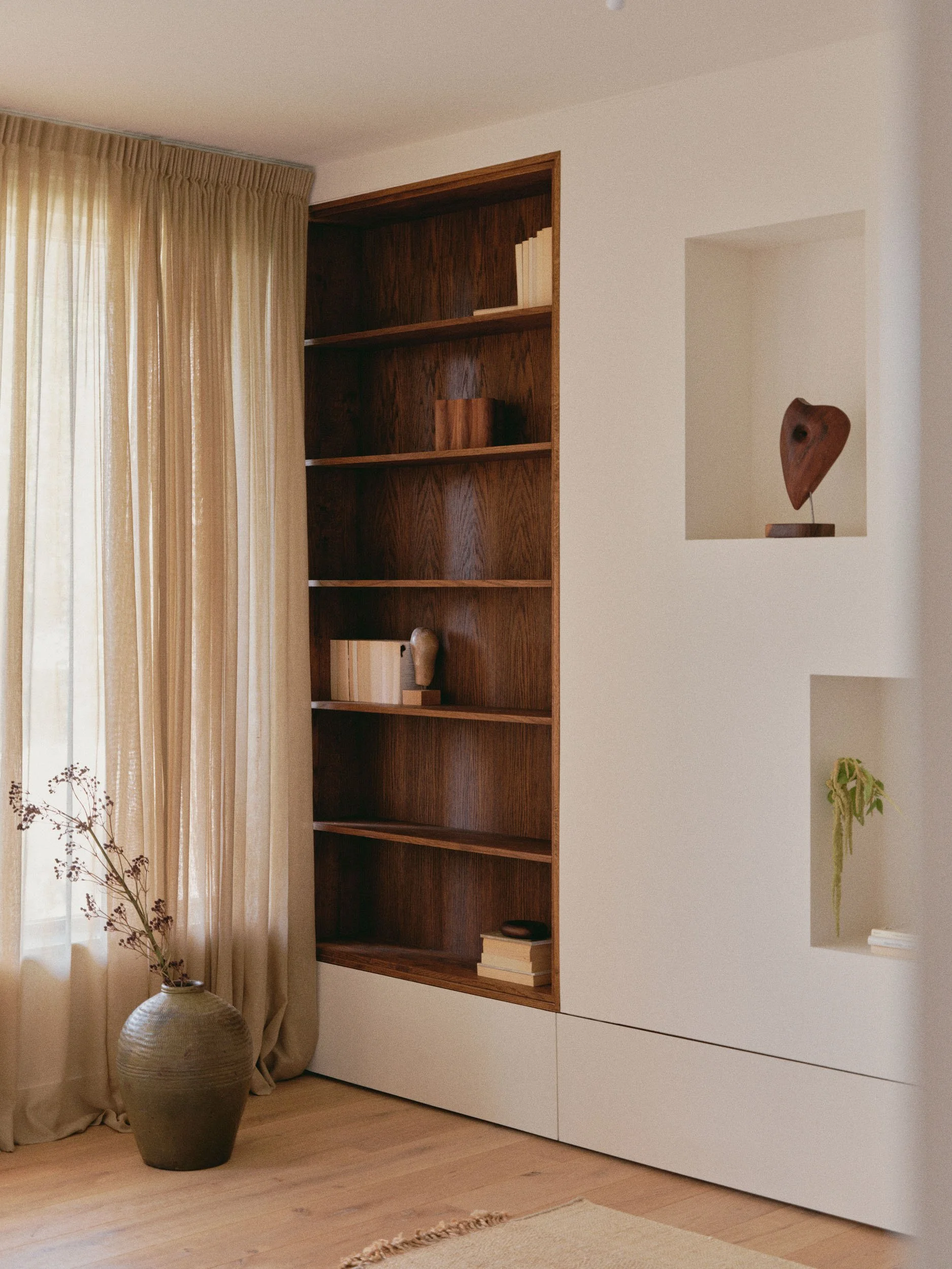 A corner of a room with beige curtains, a large ceramic vase with dried branches, a built-in wooden bookshelf with a few books and decorative items, and a white wall with open recessed shelves holding a wooden sculpture and a small plant.