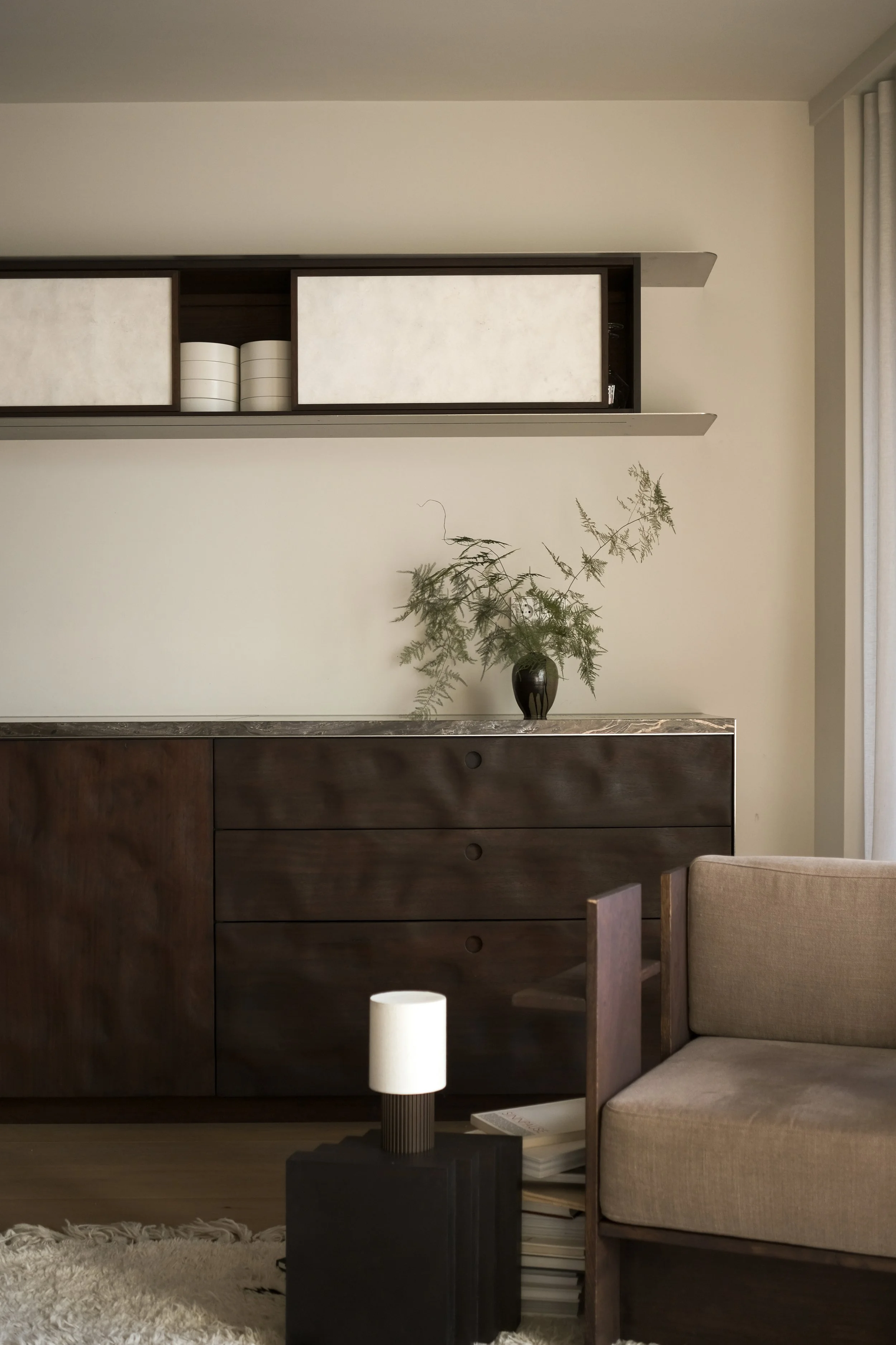Minimalist living room with a dark wooden sideboard, a beige armchair, a small black side table with a white lamp, a plant in a black vase on the sideboard, and white curtains.