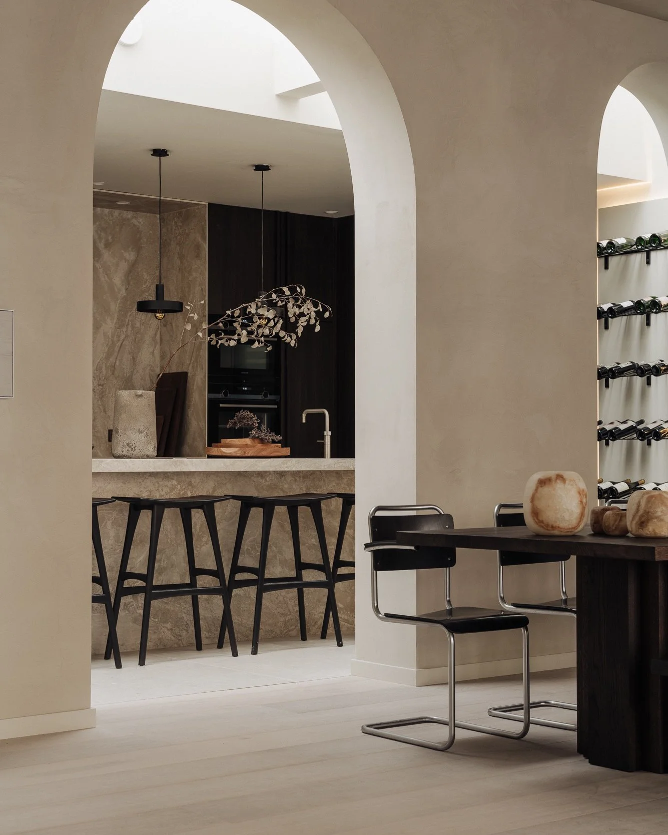 Modern kitchen and dining area with black bar stools, a dark wood table with salts, wine rack in the background, and archways with light-colored walls.
