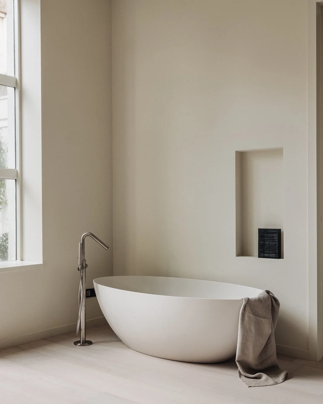 Modern bathroom with a white freestanding bathtub, a floor-mounted faucet, and a large window with natural light, with a towel draped over the edge of the tub.