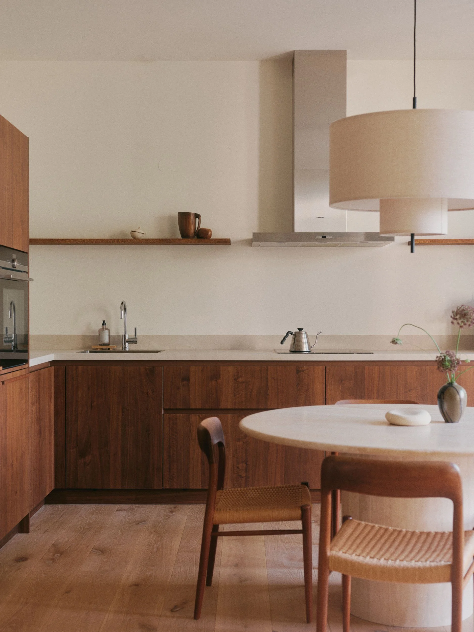 A minimalist kitchen with wooden cabinets, a beige countertop, a stainless steel range hood, and a round wooden dining table with chairs. There are a few decorative items and a vase with flowers on the table.
