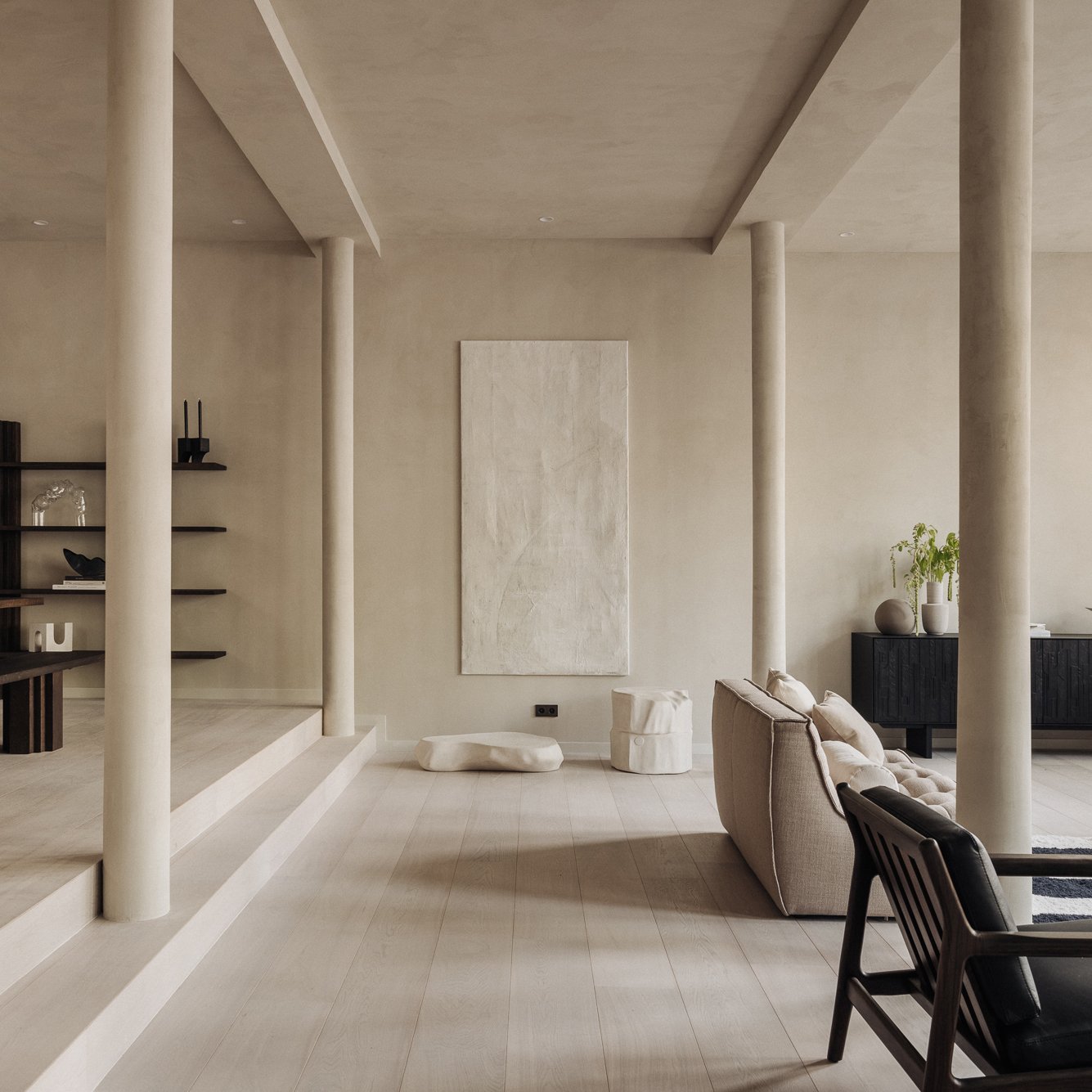 Minimalist living room with beige walls, light wood flooring, and modern decor. Features a beige sofa, black armchair, black sideboard with vases and plants, and abstract wall art.