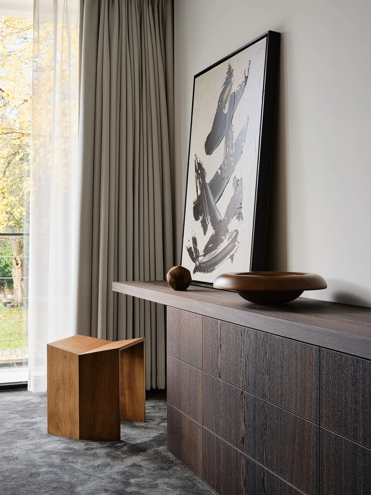 Modern interior with a wooden sideboard, a black framed abstract art piece leaning against the wall, a wooden decorative object, and a shallow bowl on the sideboard. There are curtains and a sliding glass door with a view of outdoors.
