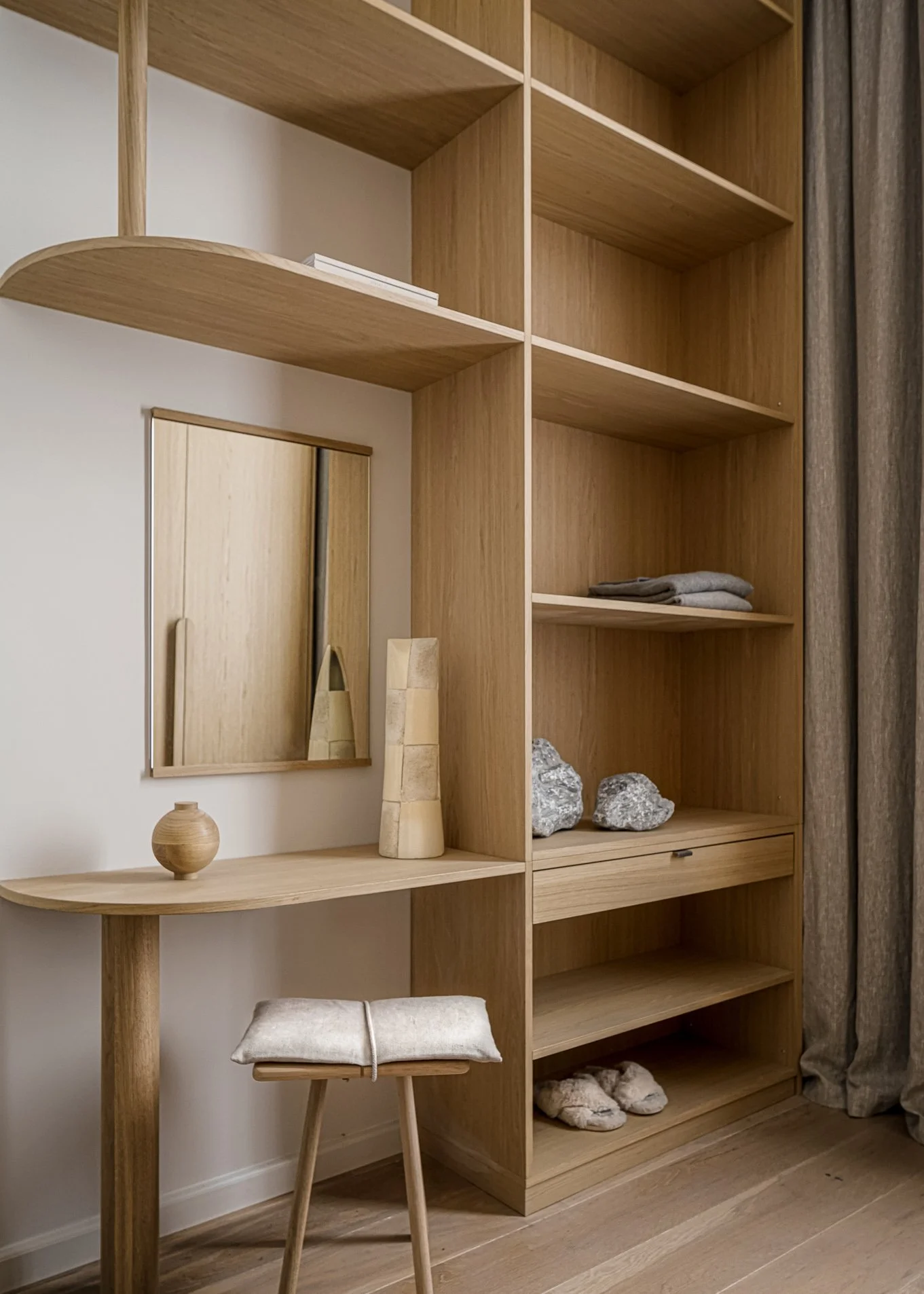 A wooden shelving unit with various empty compartments, a small desk with a stool, decorative stones, a mirror, and neatly folded towels.