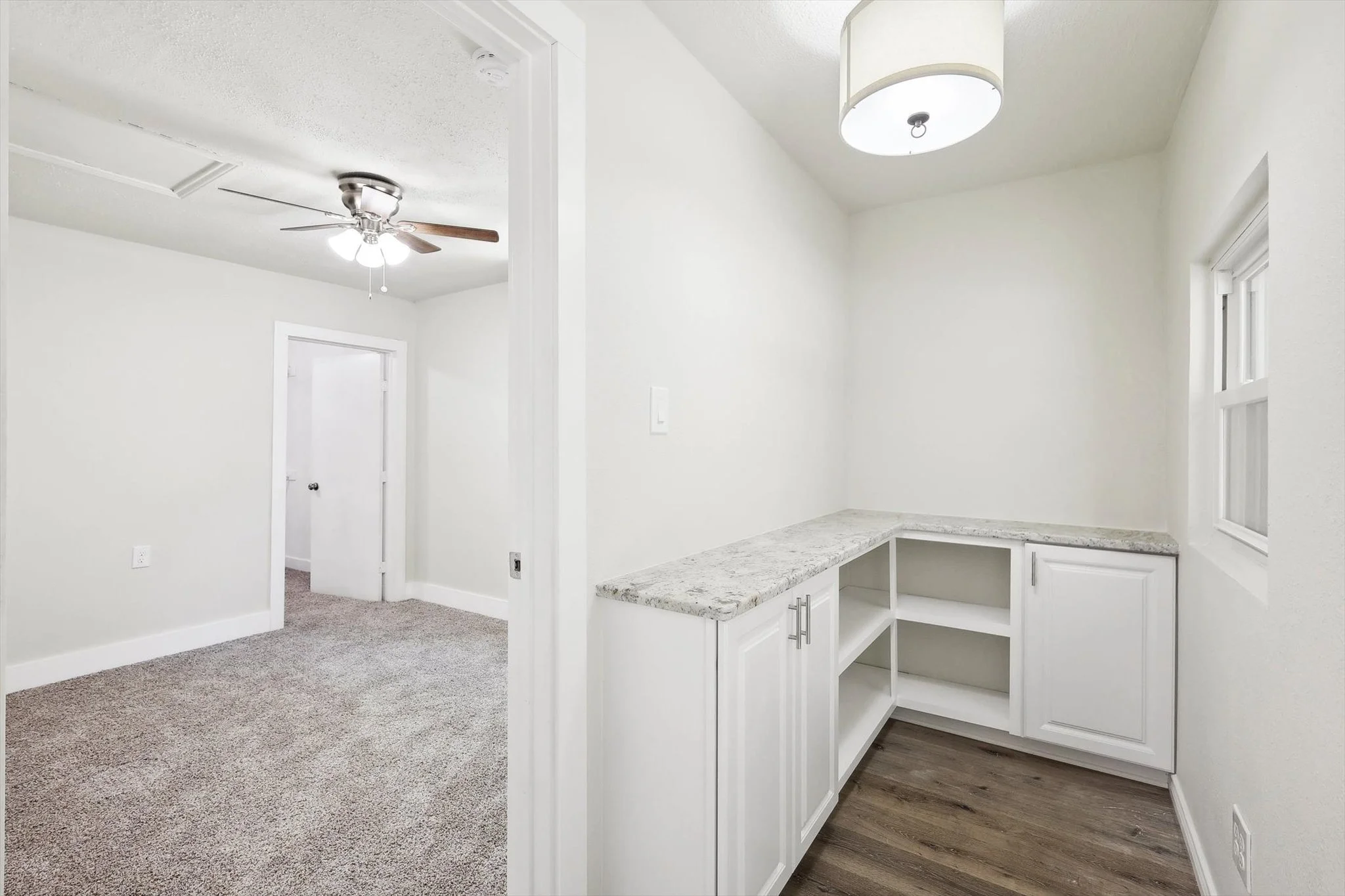 Empty kitchen nook in Euless, TX with white cabinets, granite countertop, and a small window, adjacent to a room with a ceiling fan and carpeted floor.