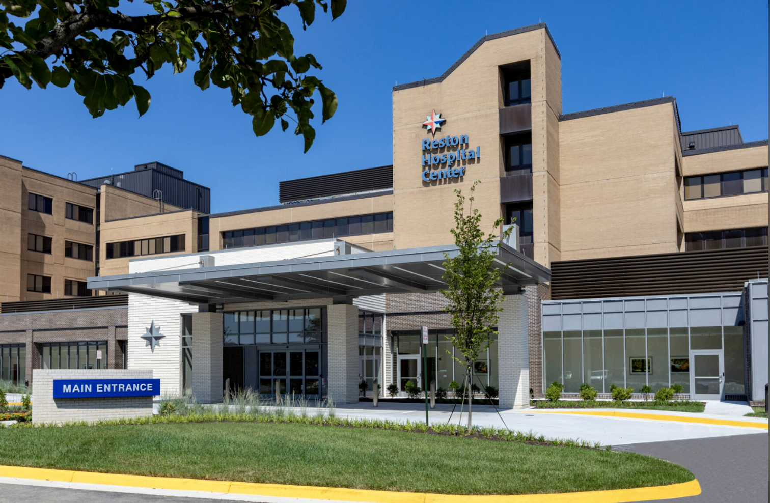 Exterior view of Reston Hospital Center, a modern medical facility with multiple levels, large windows, a main entrance marked by a blue sign, and a landscaped area in front.