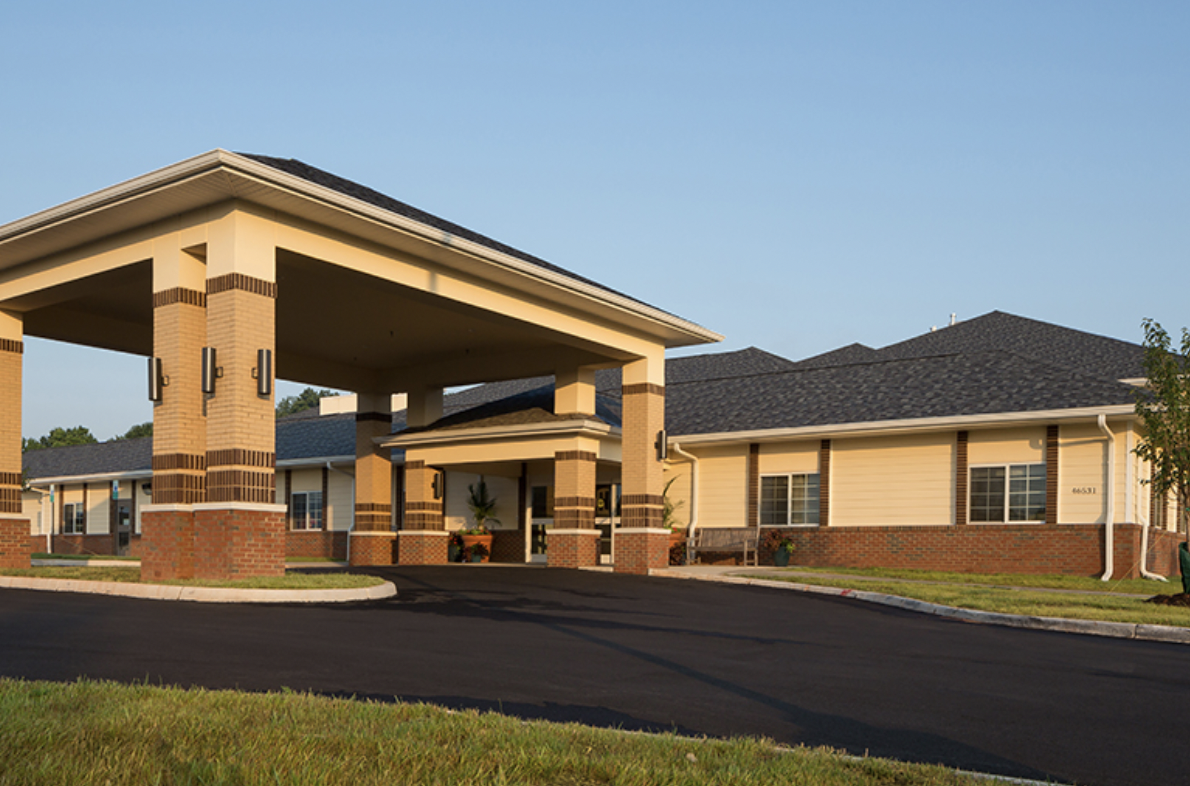 Exterior view of a modern assisted living or retirement community with a large covered entrance, brick and siding building, and well-maintained landscaping.