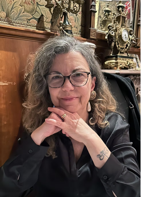 A middle-aged woman with curly gray hair and glasses, wearing earrings and a black top, sitting in a warmly decorated room with antique clocks and a tapestry in the background.
