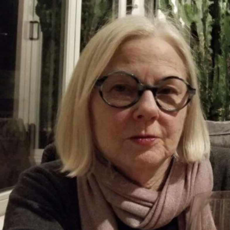 An older woman with blonde hair wearing glasses and a pink scarf. She is indoors with a background of green plants and glass windows.