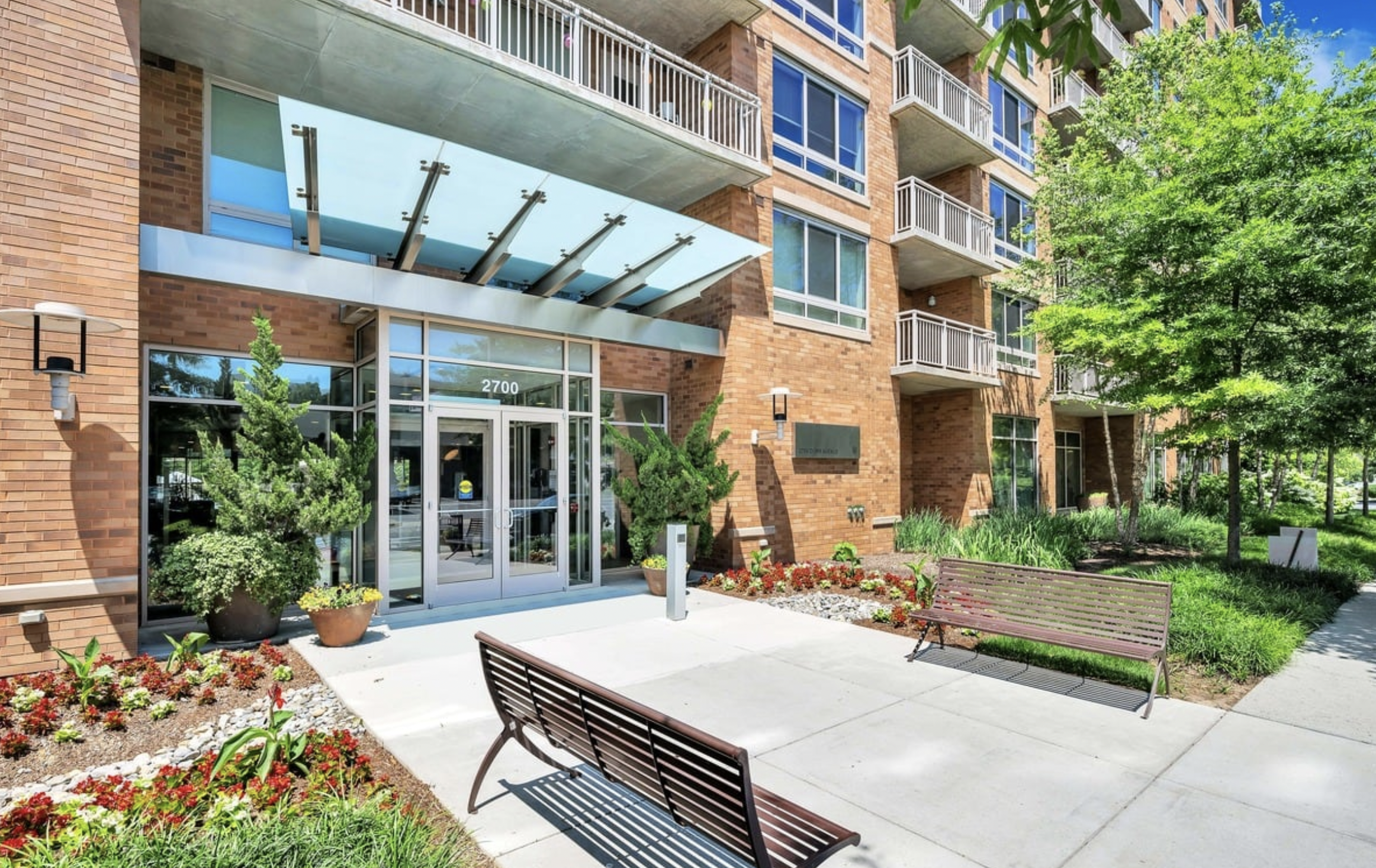 Exterior view of a modern brick apartment building with glass doors, multiple balconies, and a landscaped courtyard with benches, plants, and trees.