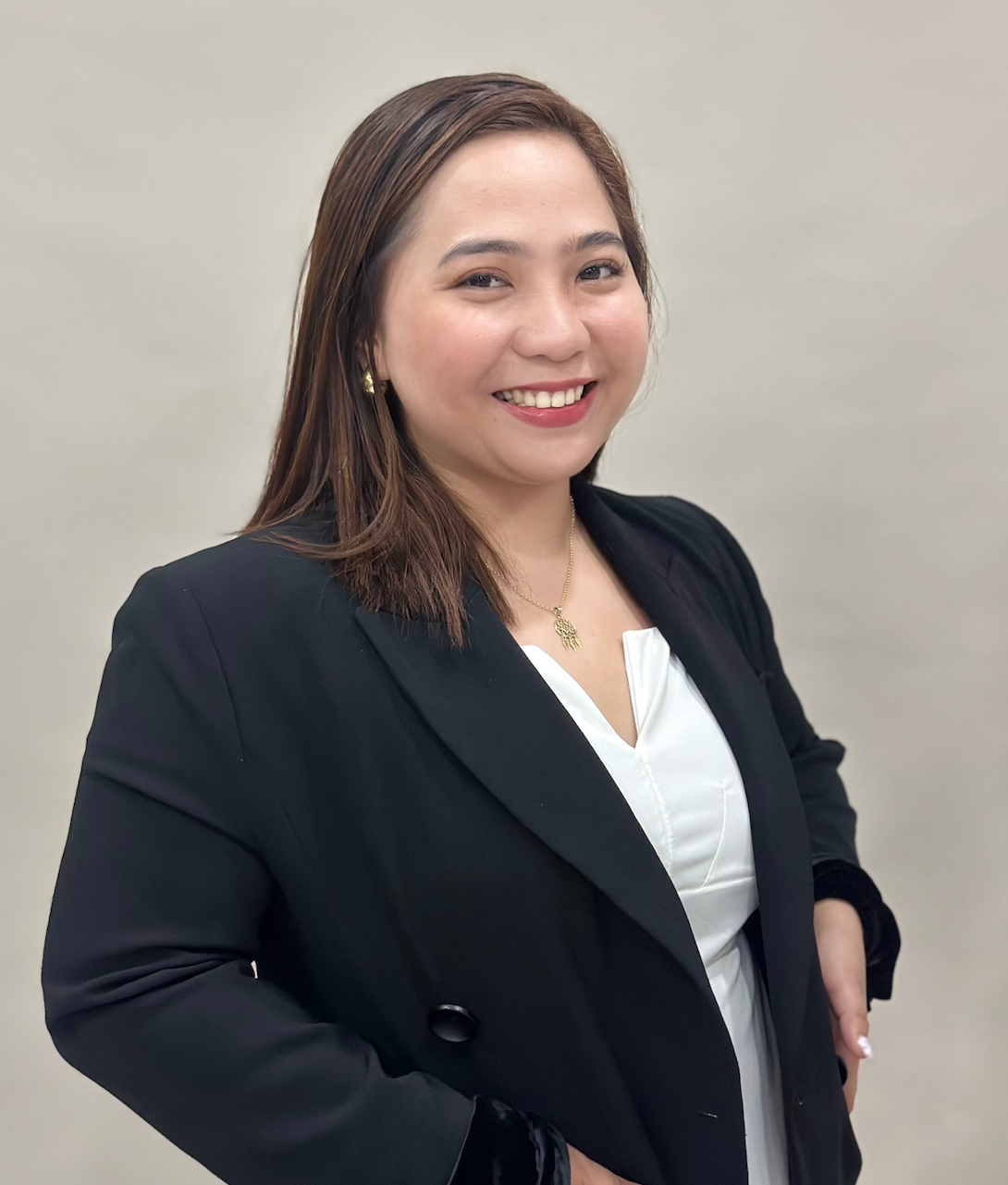 A woman with shoulder-length brown hair, smiling, wearing a black blazer, white blouse, gold earrings, and a gold necklace, standing against a plain background.