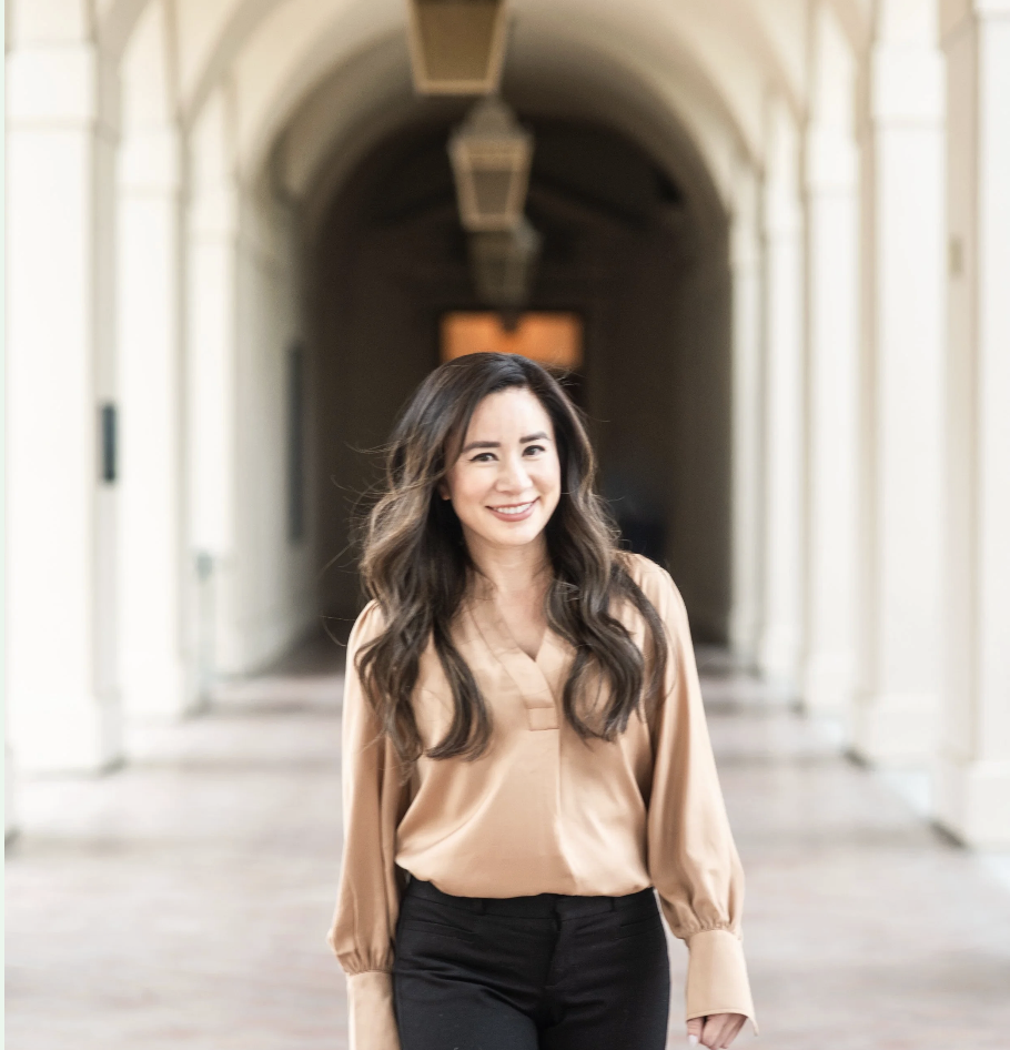A woman with long wavy hair smiling and walking through an arched hallway with hanging lights.