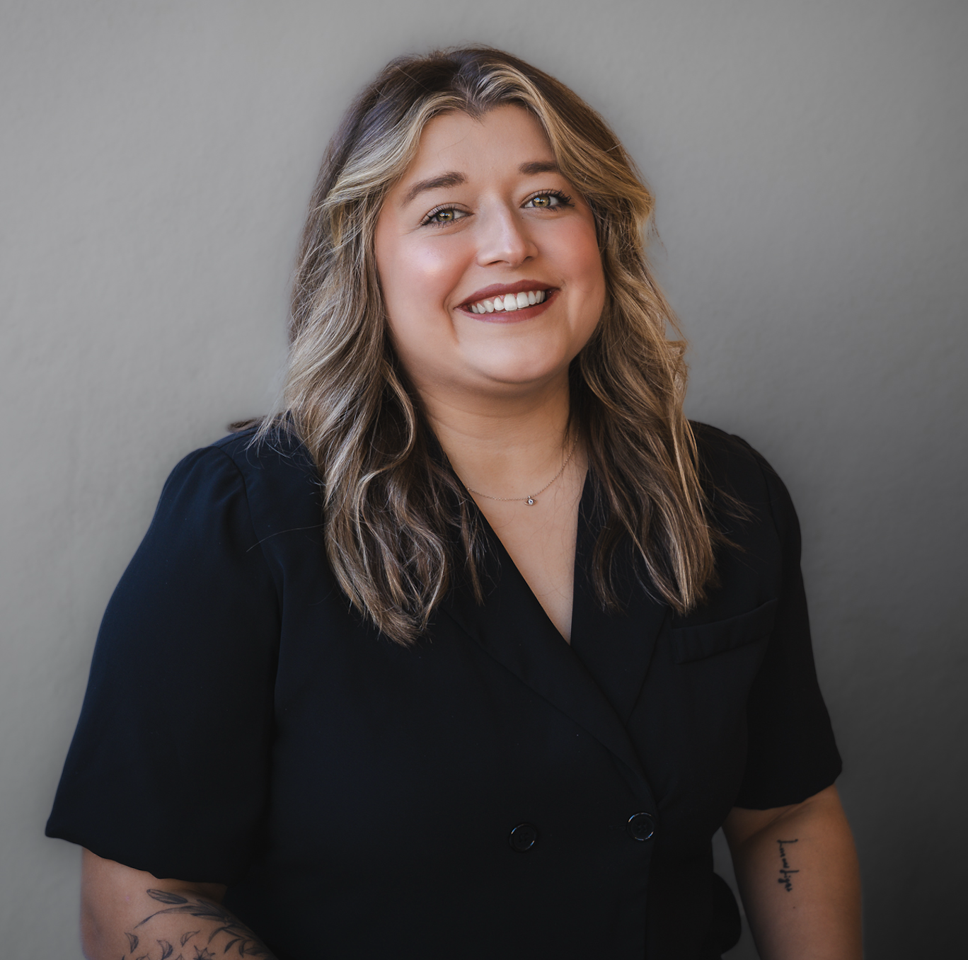Portrait of a smiling young woman with wavy, shoulder-length hair, wearing a black short-sleeve top, standing against a plain gray wall. Wearing a black top.