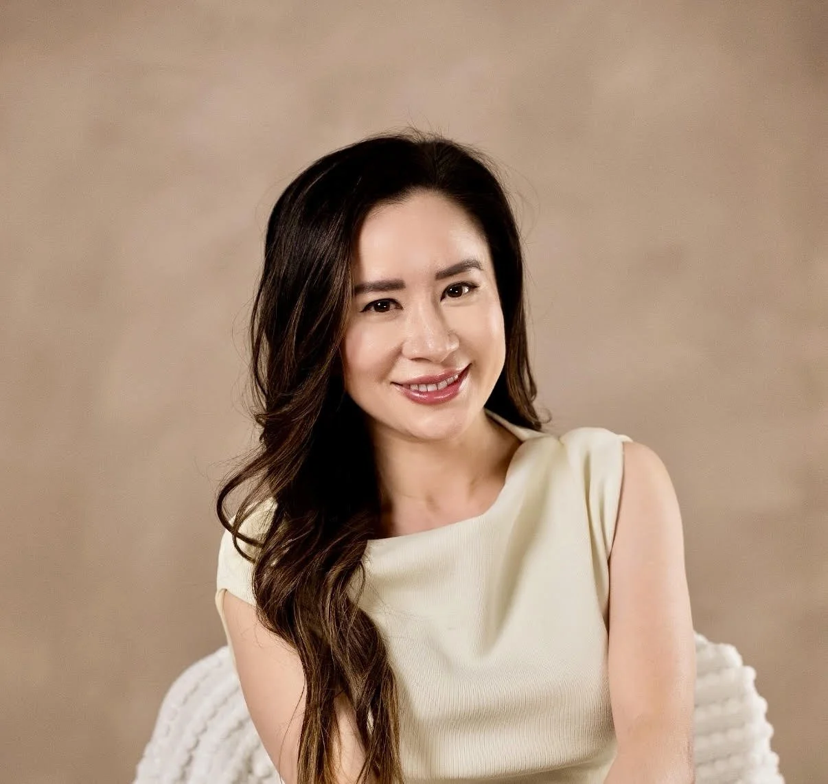 A woman with long, wavy dark hair wearing a cream-colored sleeveless top, smiling and looking at the camera against a beige background.