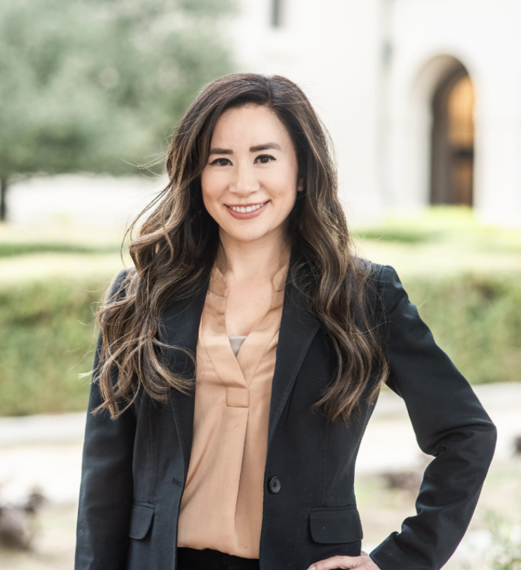 Professional woman with long wavy hair smiling outdoors, wearing a black blazer and beige blouse.