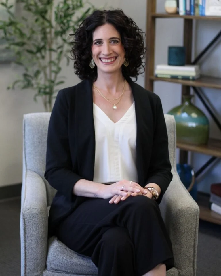 A woman sitting in a light-colored armchair, smiling, with curly dark hair, wearing a black blazer, white blouse, black pants, and jewelry, in a room with a bookshelf and plants.