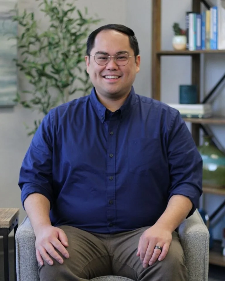 A smiling man with glasses wearing a blue button-up shirt, sitting on a light-colored upholstered chair in an office or home office setting.