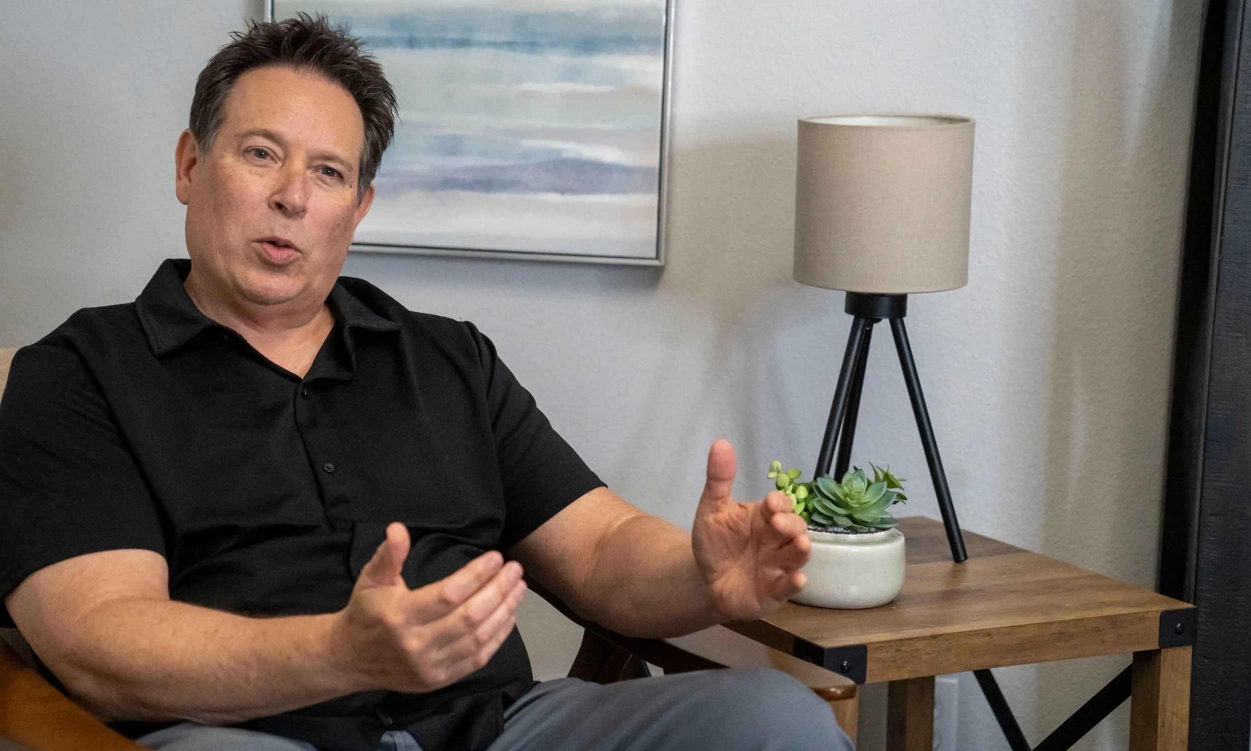 A man in a black shirt gesturing with his hands while seated near a wooden table with a small potted plant and a beige lamp, with a painting hanging on the wall behind him.