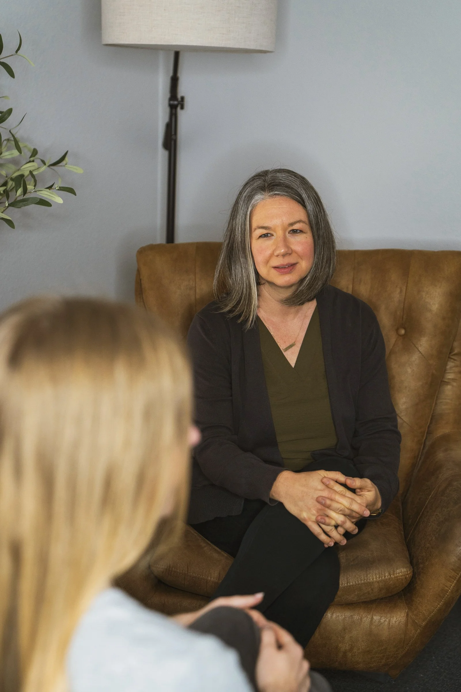 A woman with shoulder-length gray hair sitting in a brown leather armchair during a conversation. A person with long blonde hair, wearing a light-colored top, is seated opposite her, blurred in the foreground. Behind her is a gray wall, a floor lamp, and a potted plant.