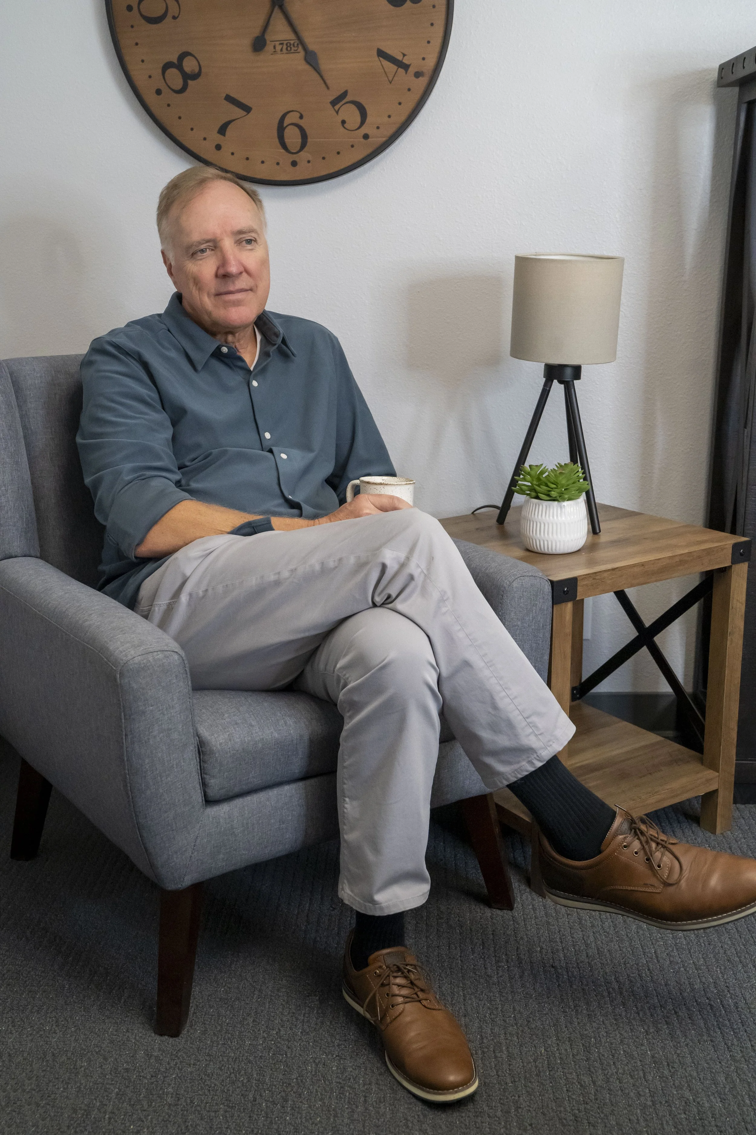 A man sitting in a gray armchair, holding a mug, in a room with a large wall clock, a wooden side table with a lamp and a potted plant.