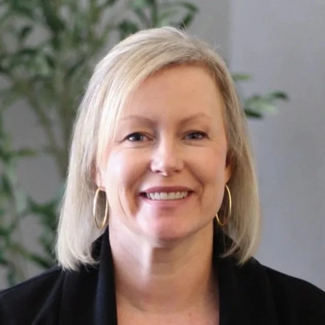 Portrait of a blonde woman smiling, wearing hoop earrings and a black outfit, with a blurred plant in the background.