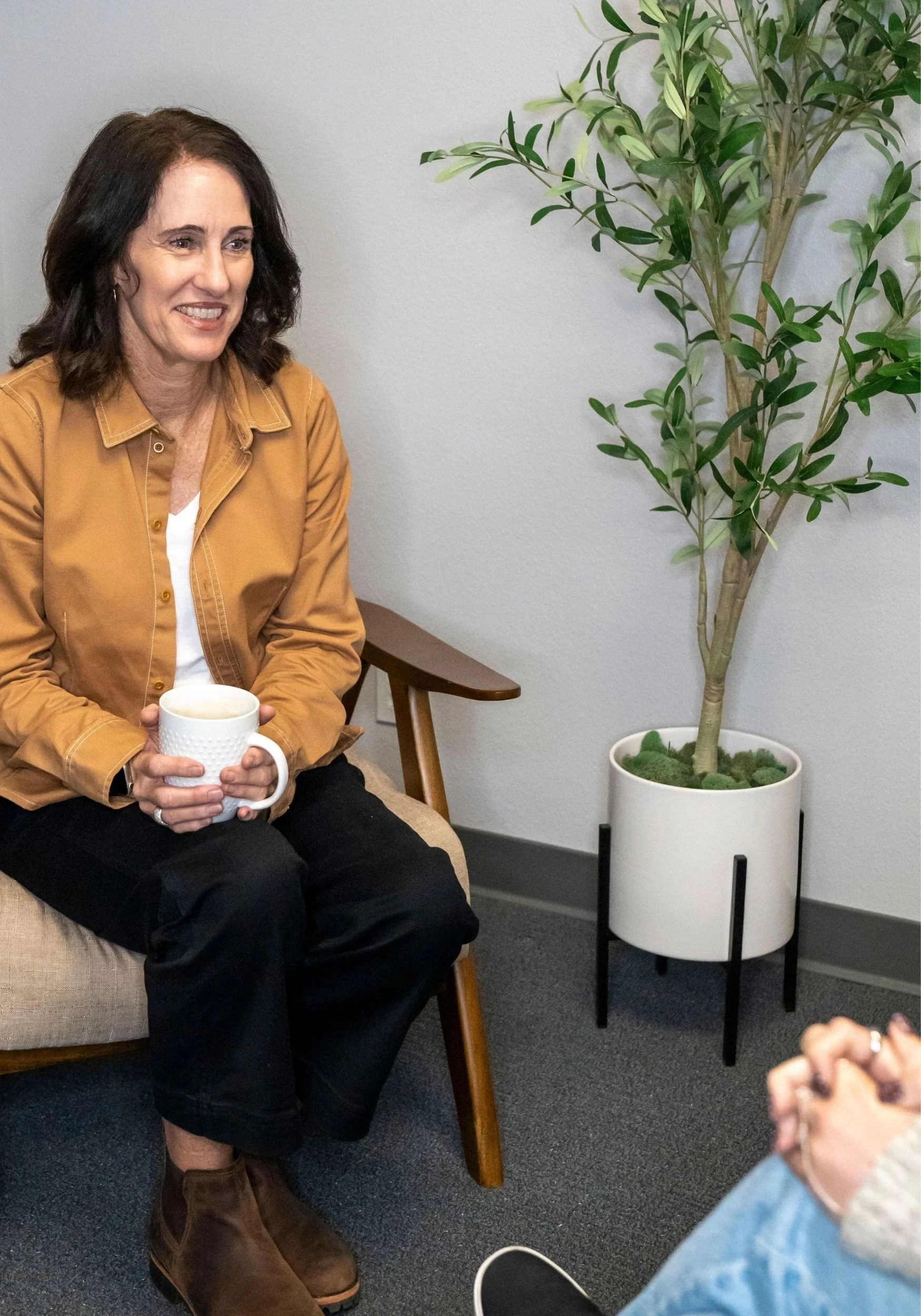 A woman sitting on a beige couch with a white mug in her hands, wearing a brown jacket and black pants, in a room with a plant in a white pot nearby.