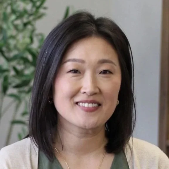 Smiling woman with shoulder-length black hair, wearing a beige cardigan over a green top, standing indoors near a plant.