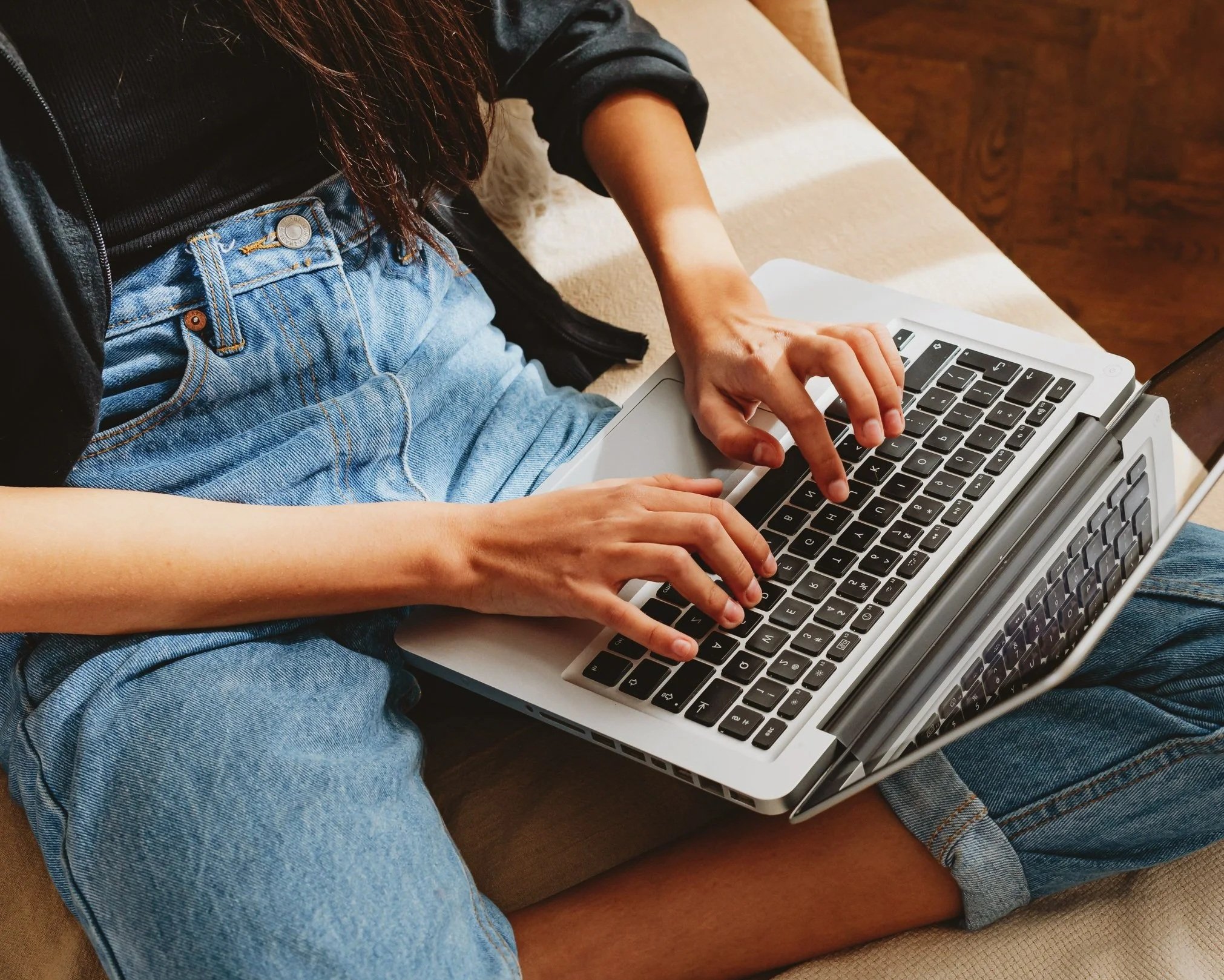 Person sitting on a couch using a laptop, wearing a black jacket, black top, and blue jeans.