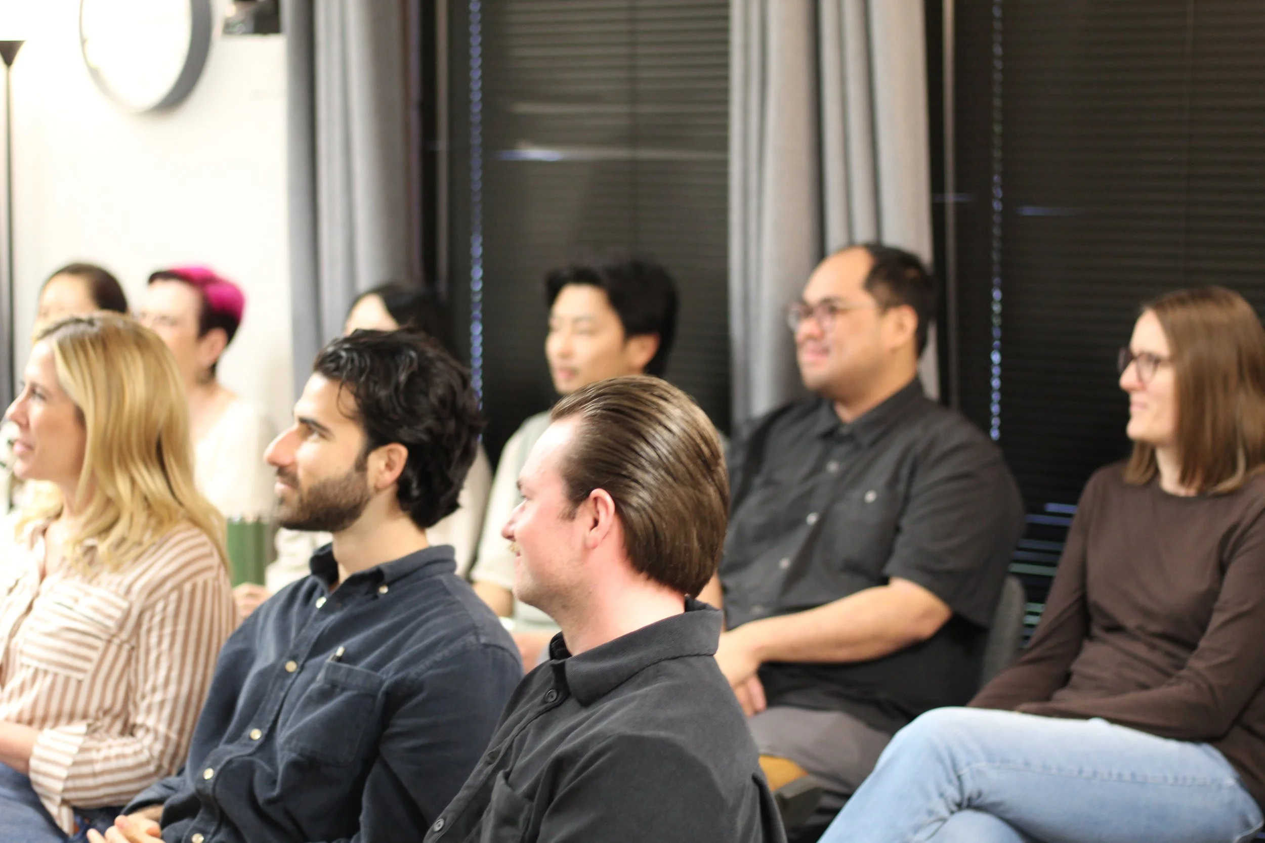 People sitting attentively at an indoor event or seminar, facing forward, with a mix of men and women of different ethnicities.
