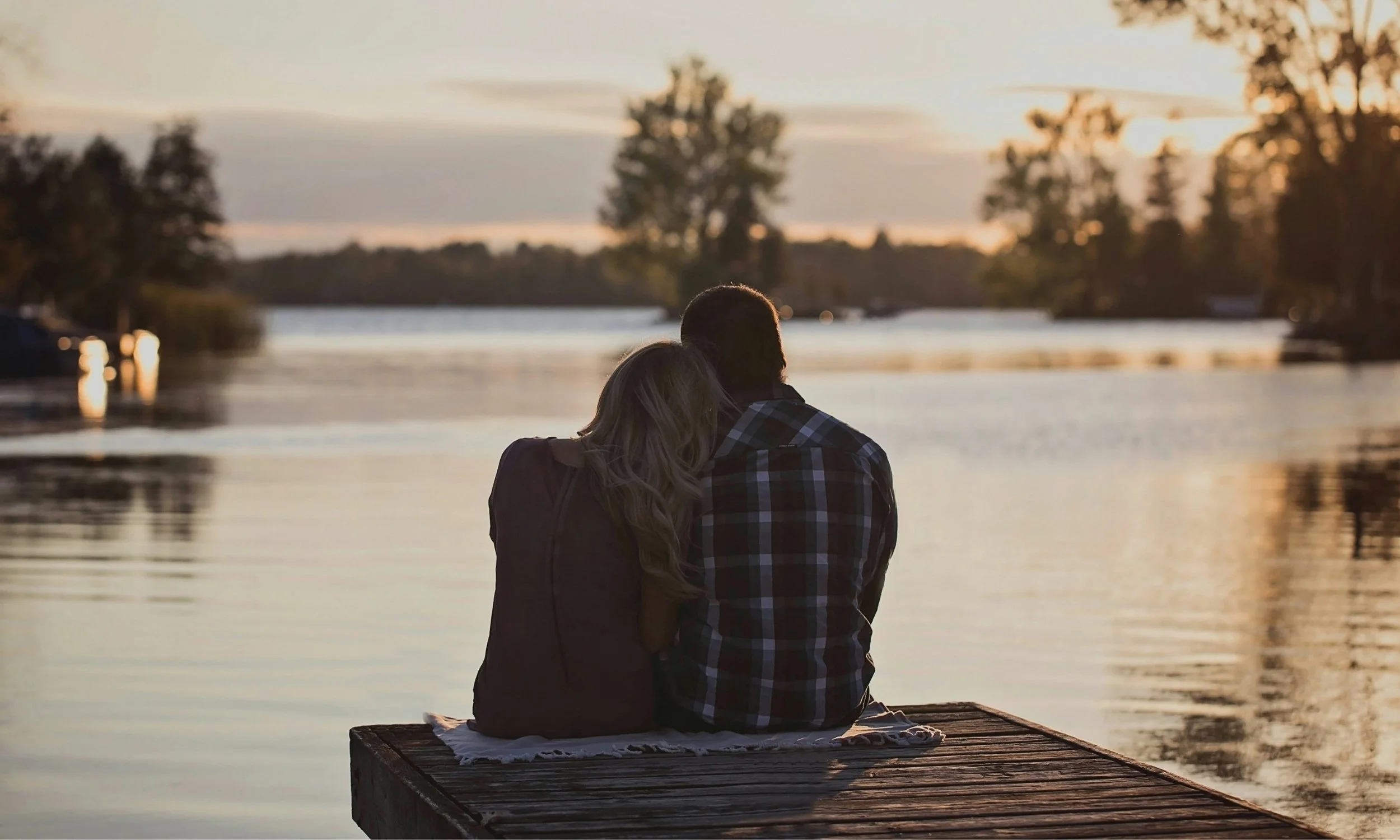 A couple sits on a dock by a body of water during sunset, with trees in the background.