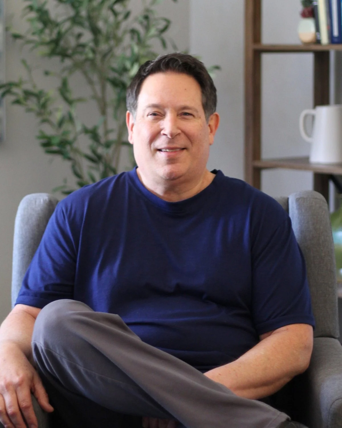 A man with dark hair, wearing a navy blue T-shirt and gray pants, sitting on a gray chair in a room with a wooden bookshelf, a large potted plant, and a white mug on the shelf.