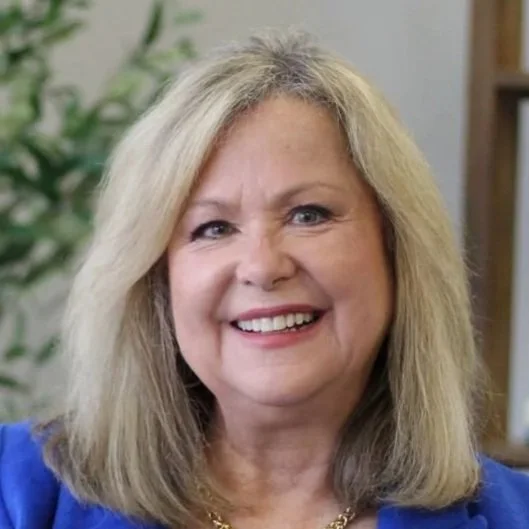 Smiling woman with shoulder-length blonde hair, wearing a blue blazer and a gold necklace, in a room with a plant and wooden shelves in the background.