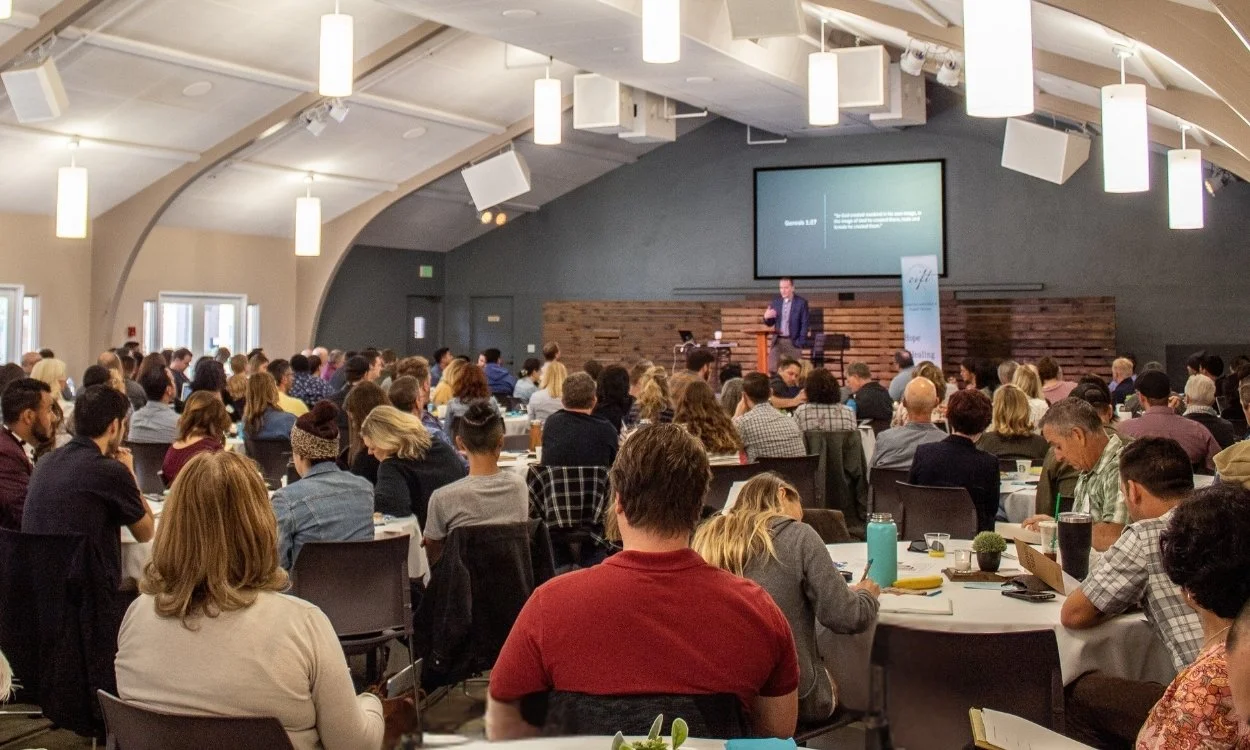 A large conference room filled with people attending a presentation. The speaker is on stage with a large screen behind them, and some attendees are taking notes or looking at their devices.