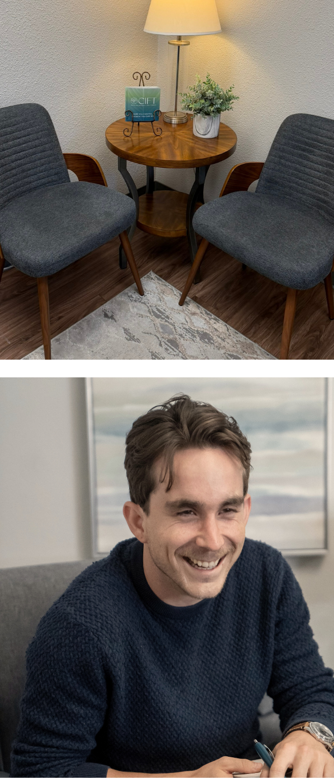 A cozy waiting area with a round wooden table, two gray upholstered chairs, a lamp, a plant, and a candle holder on the table; a smiling man in a navy sweater sitting at a desk.