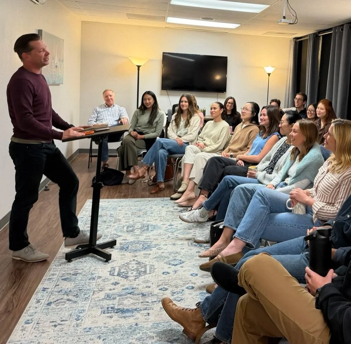 A man giving a presentation to a group of smiling women and men in a small meeting room with a flat-screen TV on the wall and floor lamps, seated on chairs, listening attentively.