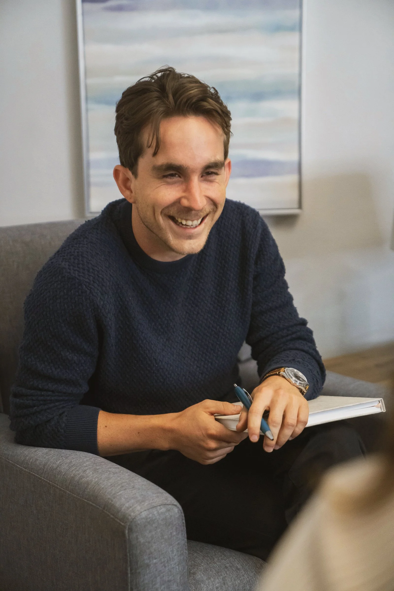 A smiling man with short brown hair, wearing a navy blue sweater, is sitting on a gray sofa with a notebook and pen in his hands. Behind him, there is an abstract painting with blue, white, and gray stripes.
