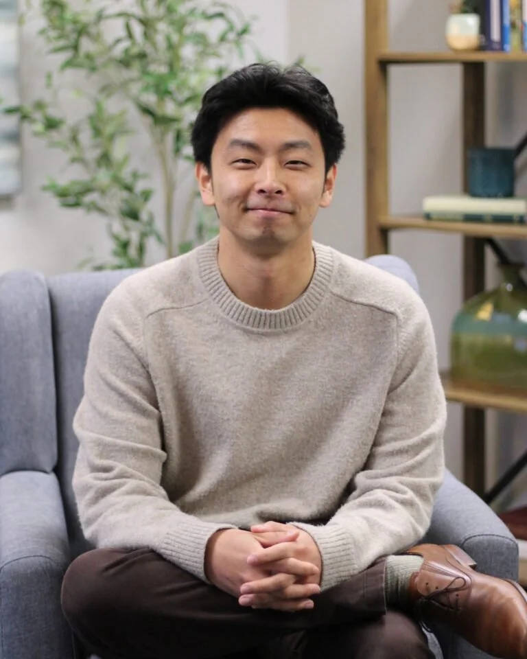 A young man with short black hair and a light smile, sitting on a gray couch in a cozy room with a bookshelf and green plant in the background.