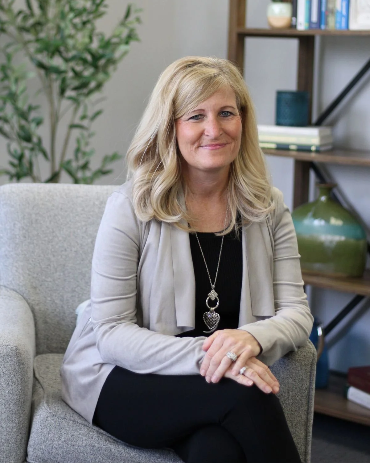 A woman with blonde hair sitting on a beige armchair in an office or library setting, smiling and looking at the camera. She is wearing a black top, a beige cardigan, a large heart-shaped pendant necklace, and rings, with a bookshelf and decorative items in the background.