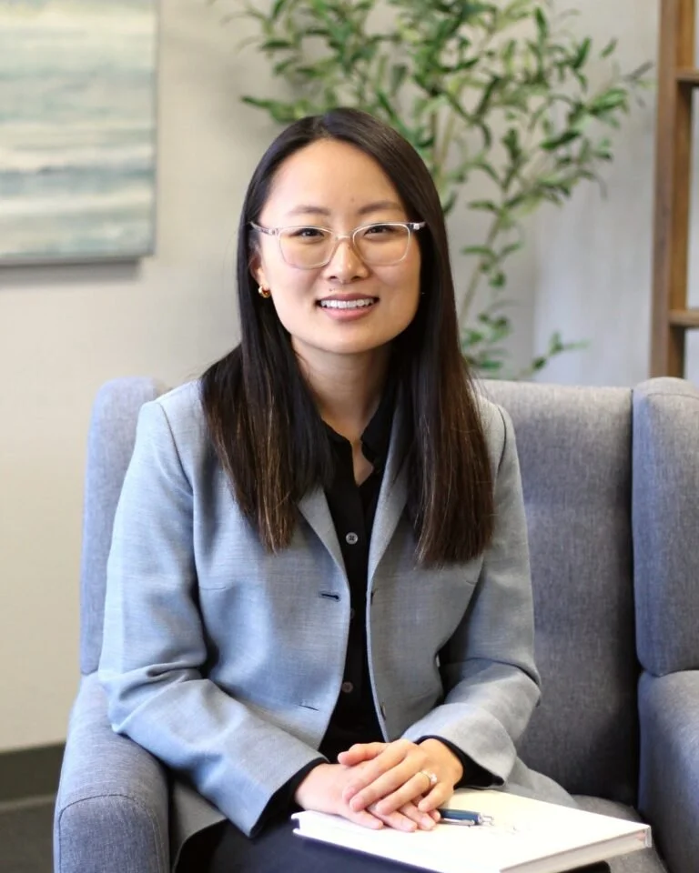 A professional woman with glasses and long dark hair, sitting on a gray armchair, wearing a gray blazer over a black top, smiling, with a notepad and pen on her lap, in a modern office setting with a plant and artwork in the background.