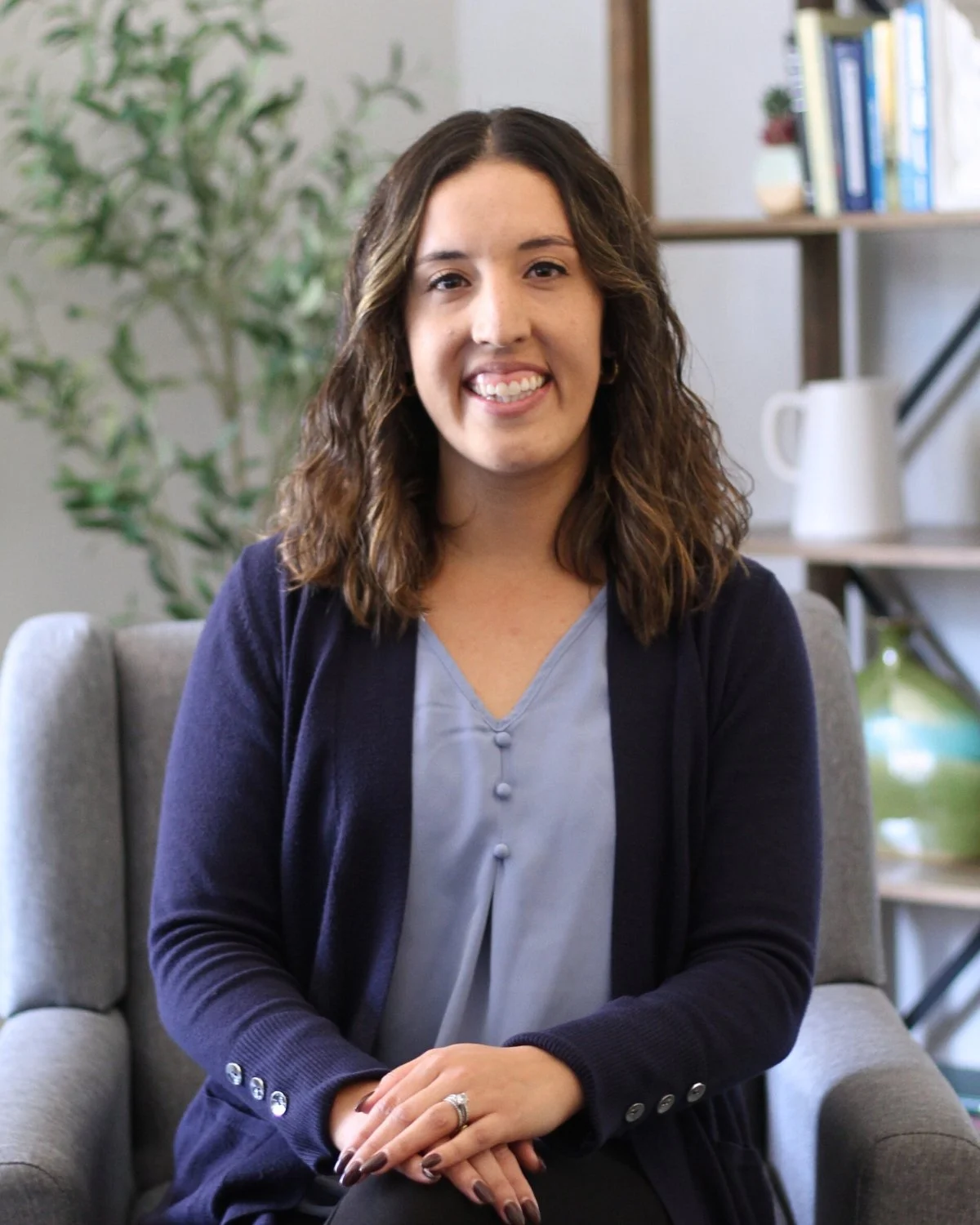 A woman with shoulder-length curly brown hair, smiling, seated on a gray armchair in an indoor setting with a bookcase and potted plant in the background.