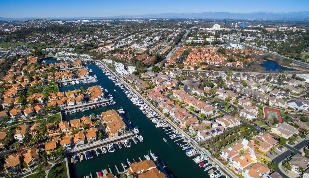 Aerial view of a residential neighborhood with a waterway surrounded by houses with orange rooftops and docks, green trees, and a tennis court. In the background are more houses, roads, and distant mountains under a clear blue sky.