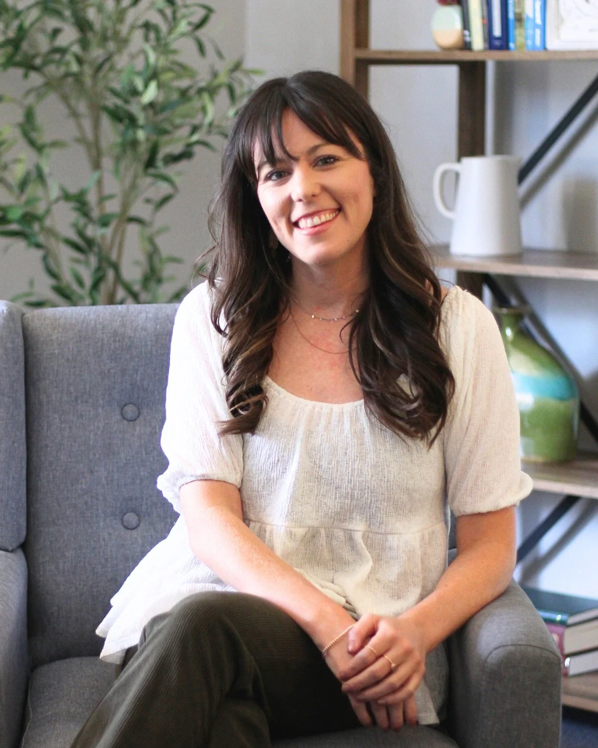 A woman with long dark hair, sitting on a gray couch, smiling in a cozy, well-decorated room with a bookshelf, a plant, and decorative items in the background.
