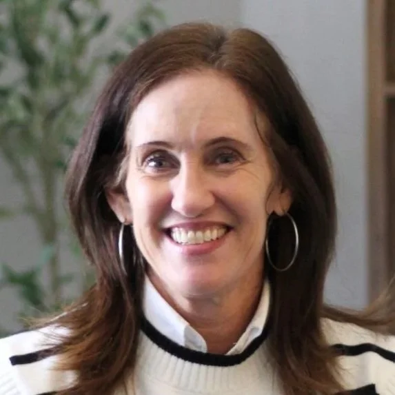 A woman with shoulder-length brown hair, wearing hoop earrings and a white sweater with black stripes, smiling indoors with a blurred plant in the background.
