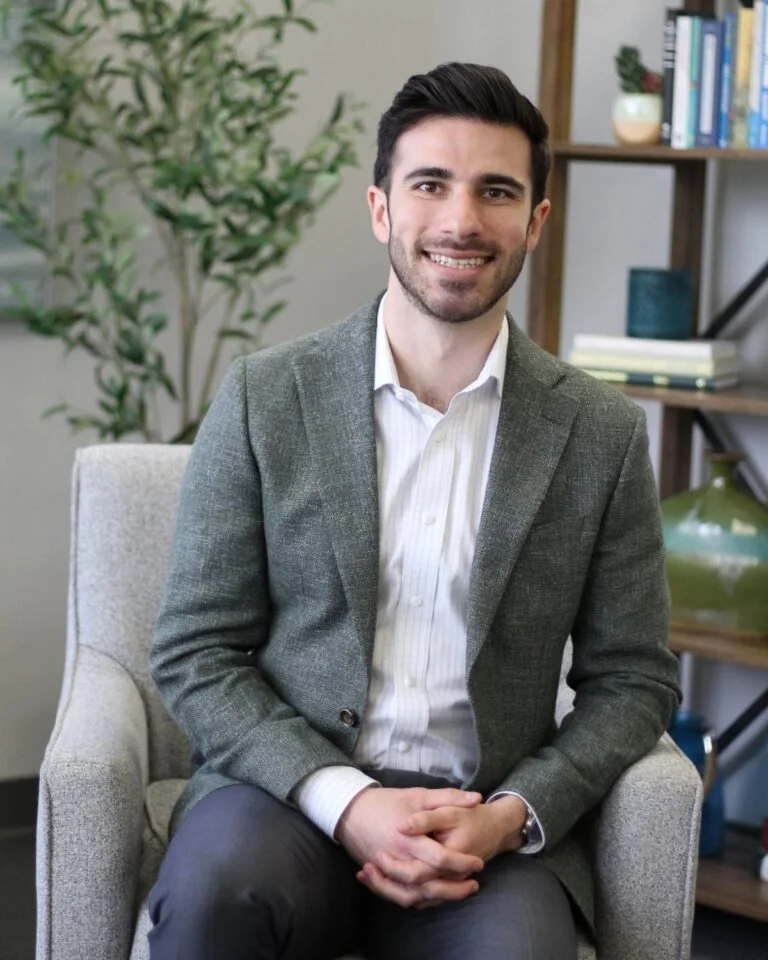 A man with dark hair, beard, dressed in a gray blazer and white shirt, sitting on a beige armchair in a room with bookcase, green plant, and decorative items.