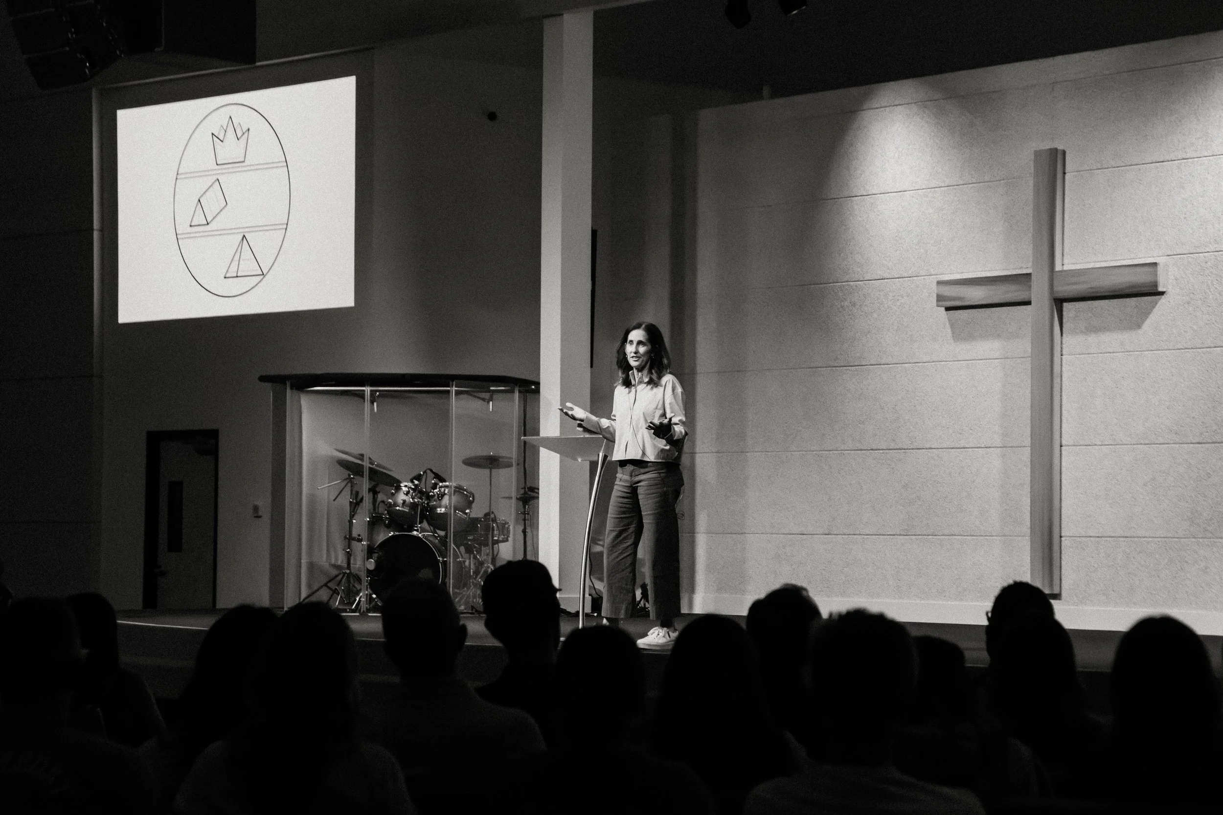 A woman giving a speech on stage at church, with a cross mounted on the wall to her right and a drum set behind her. A projected image showing geometric shapes inside an oval is on the wall to her left.