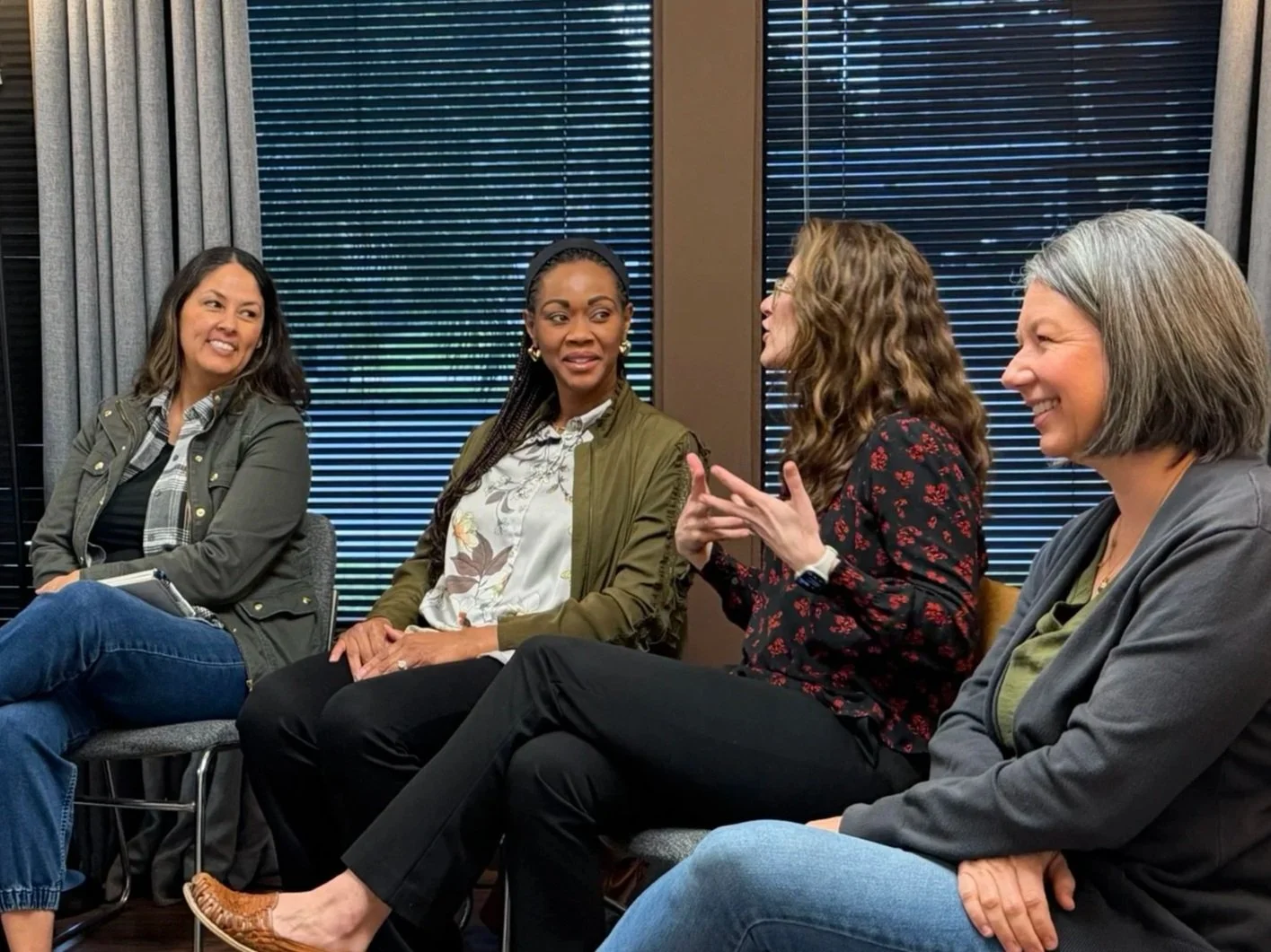 Four women sitting in a panel discussion, engaged in conversation, with black blinds and curtains in the background.