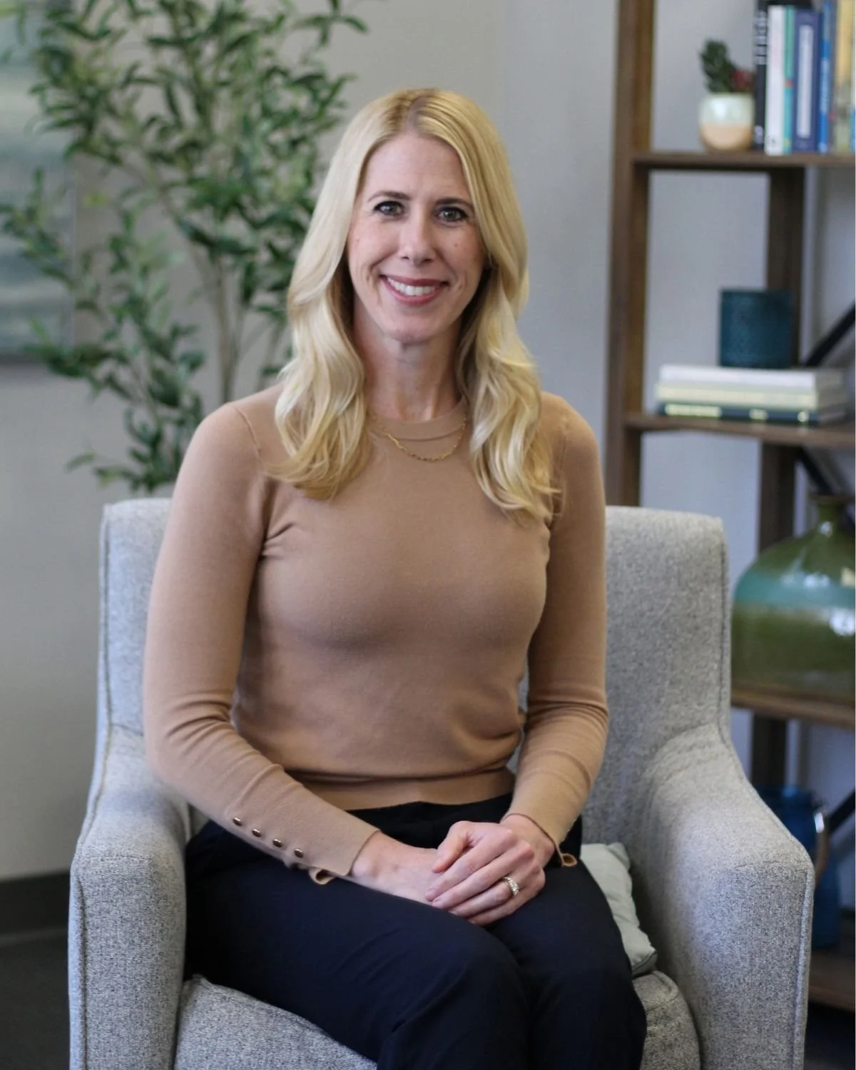 A smiling woman with blonde hair sitting in a light gray armchair in an office, with bookshelves and a plant in the background.