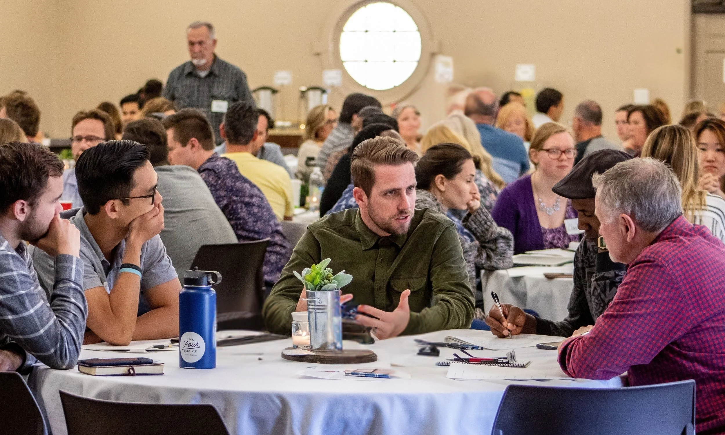 A diverse group of people seated at round tables in a conference room, engaging in discussion during a meeting or workshop, with some people taking notes and a man in the background standing near a window.