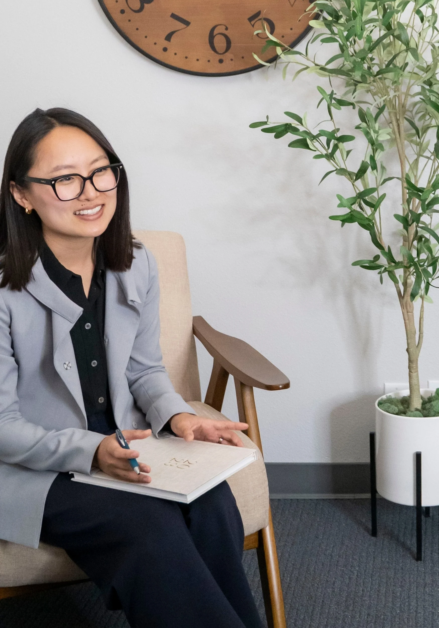 A woman with glasses and dark hair smiling, sitting on a beige chair with wooden armrests, holding a pen and notebook. There is a large clock on the wall behind her and a potted plant on the right.